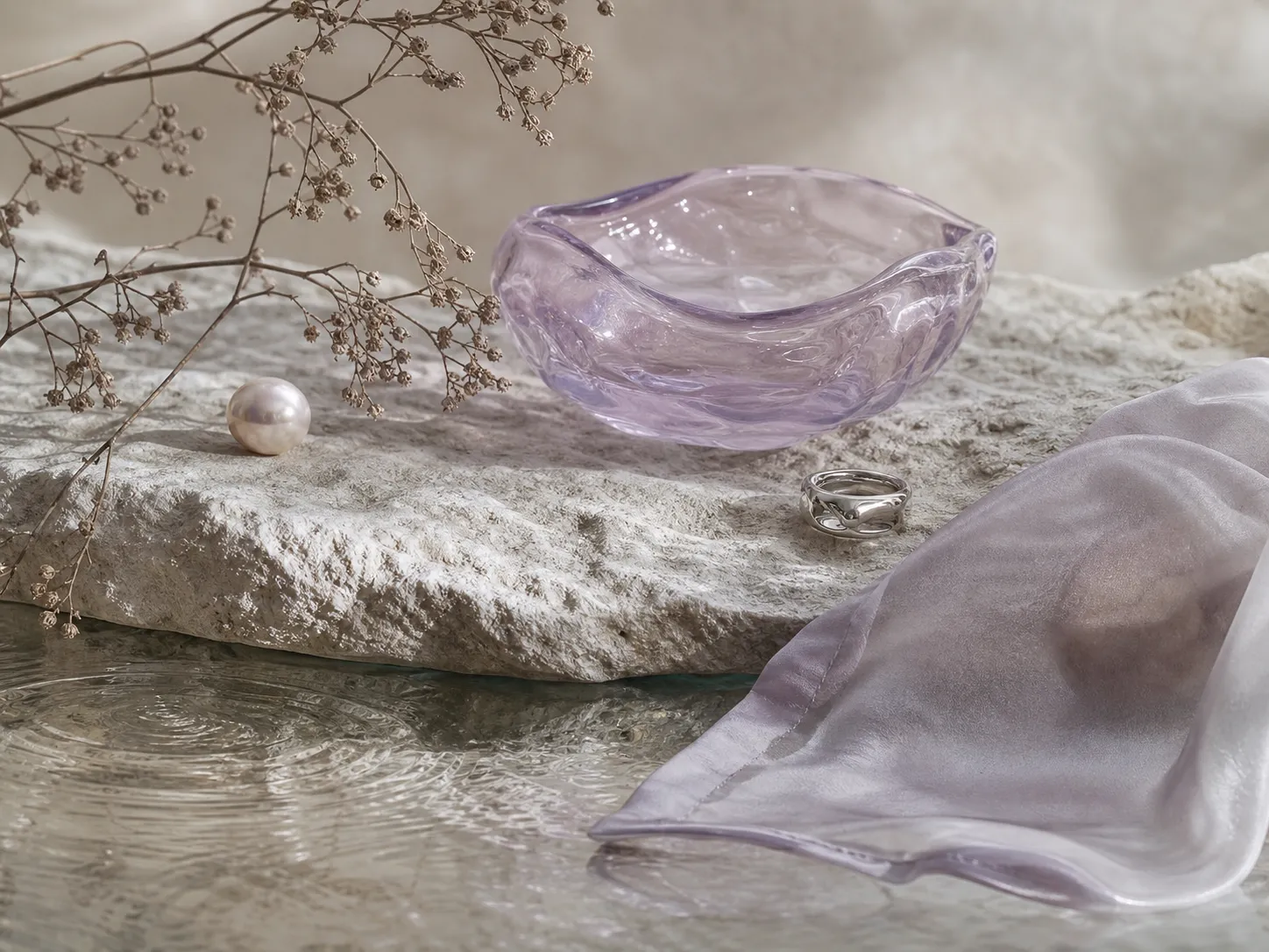 A lilac glass bowl, pearl, silver ring, branch, silk, stone, and water in a close object study