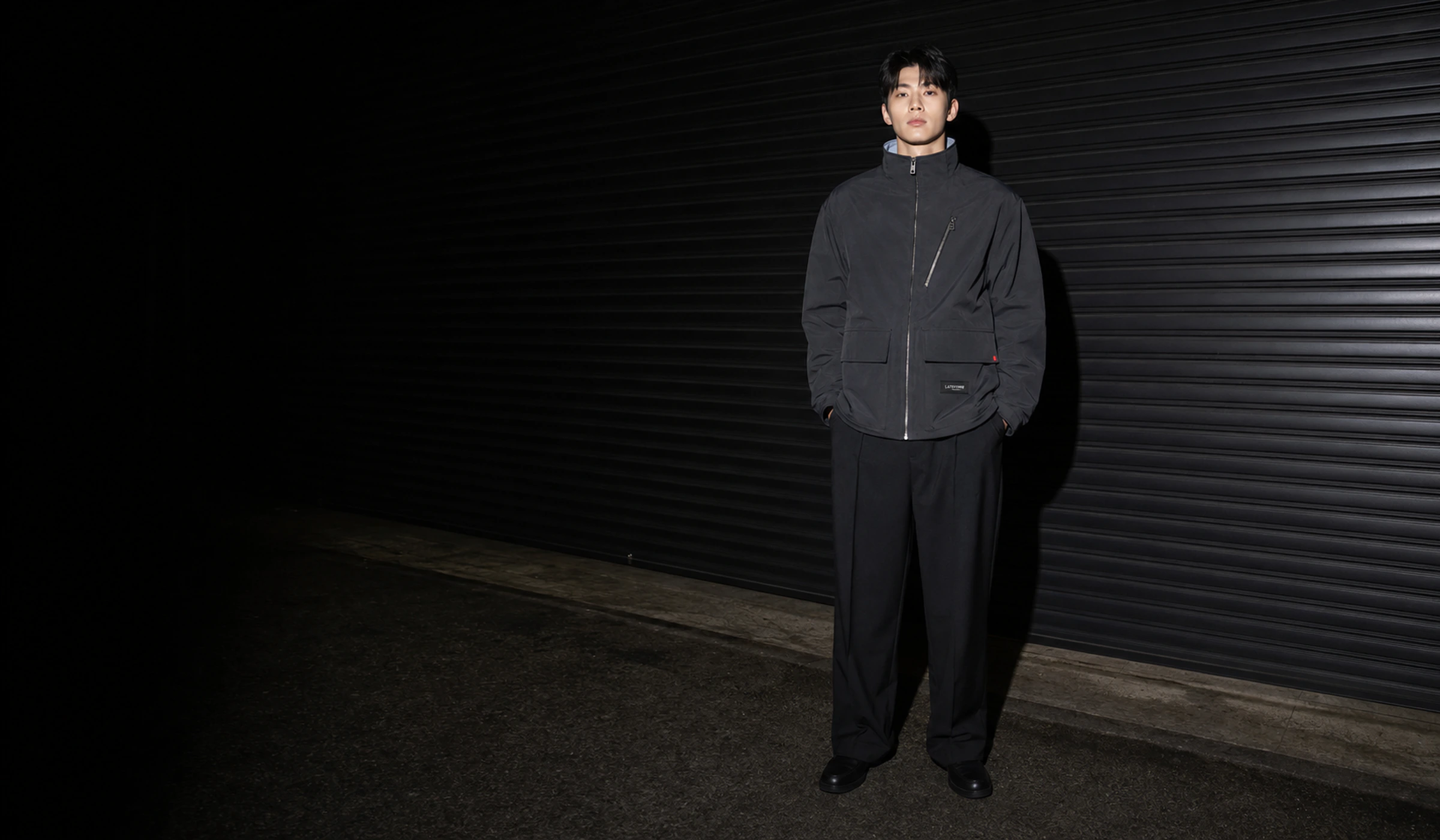 어두운 셔터 앞에서 LATEFORM RELAY JACKET을 착용한 모델